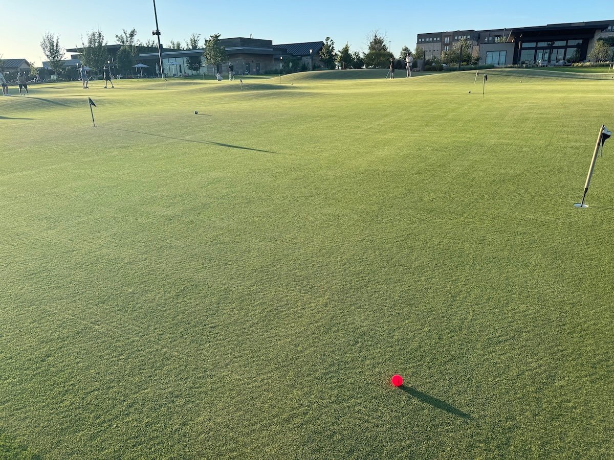 Red golf ball on a large putting green, with holes in the distance.