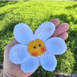 Flor em croché com pétalas brancas, centro amarelo sorridente e pormenores rosa, apoiada numa mão.