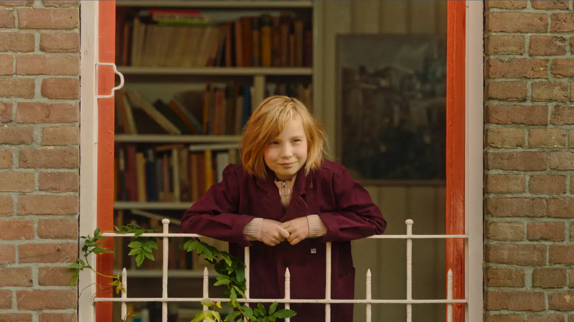 Young girl with red hair and a maroon jacket standing by a white fence in front of a bookshelf, smil.