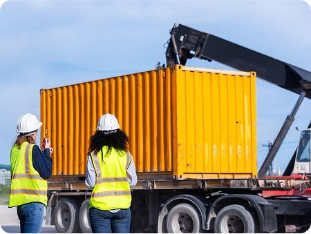 Two people in high-visibility clothing watching a shipping container being loaded onto a truck by crane