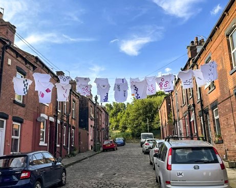 Art installation. A row of t-shirts, with art work emblazoned on them, are hanging high on a washing line between two back-to-back houses.