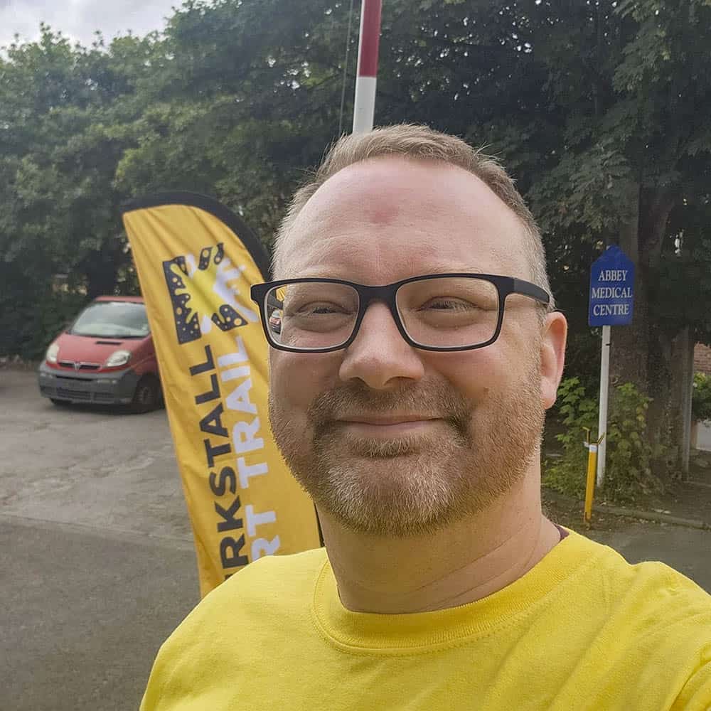Jim Corah wears a yellow t-shirt. He has short hair and wears glasses. He stands in front of a Kirkstall Art Trail banner