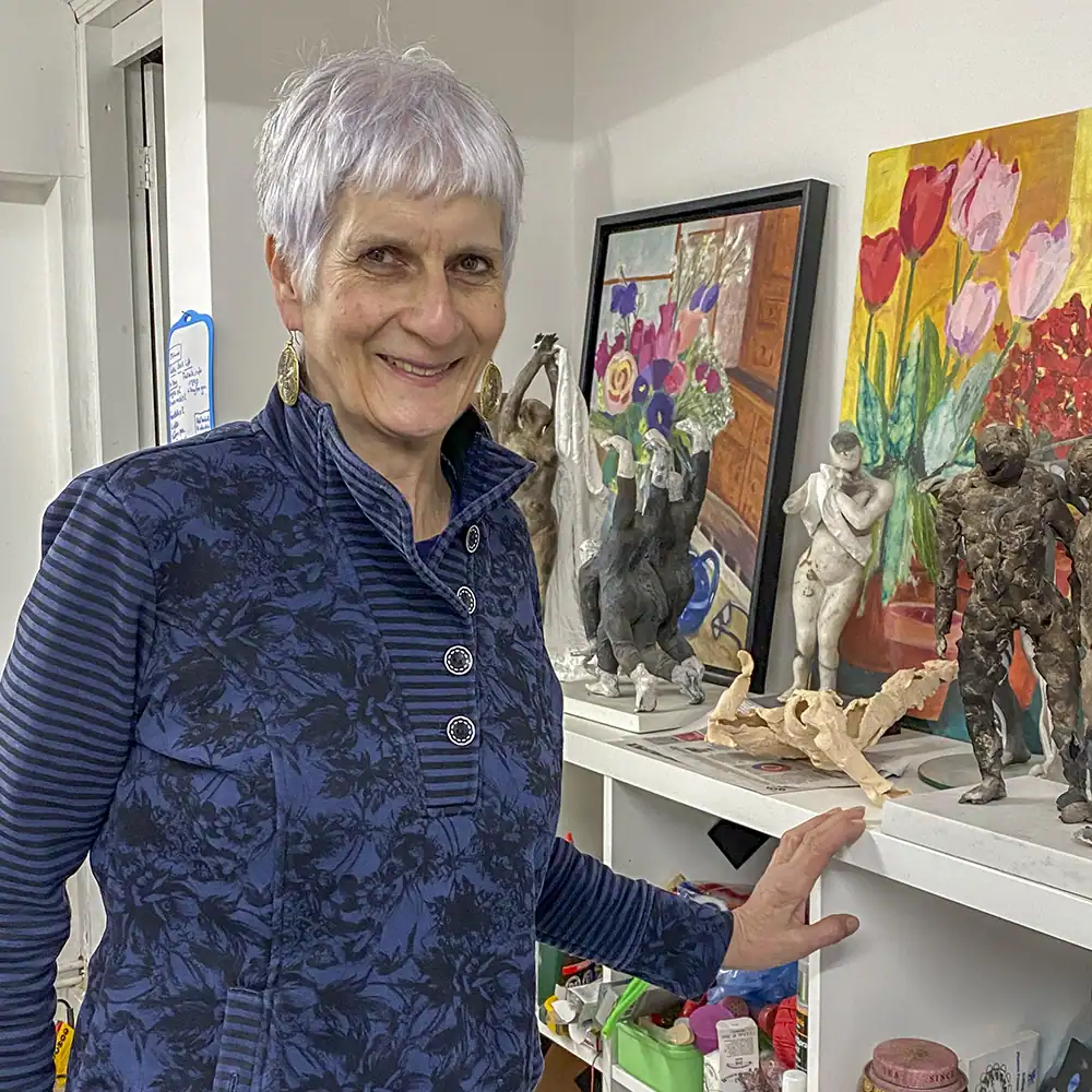 A smiling Mandy Long stands next to a shelf adorned with her sculptures and paintings