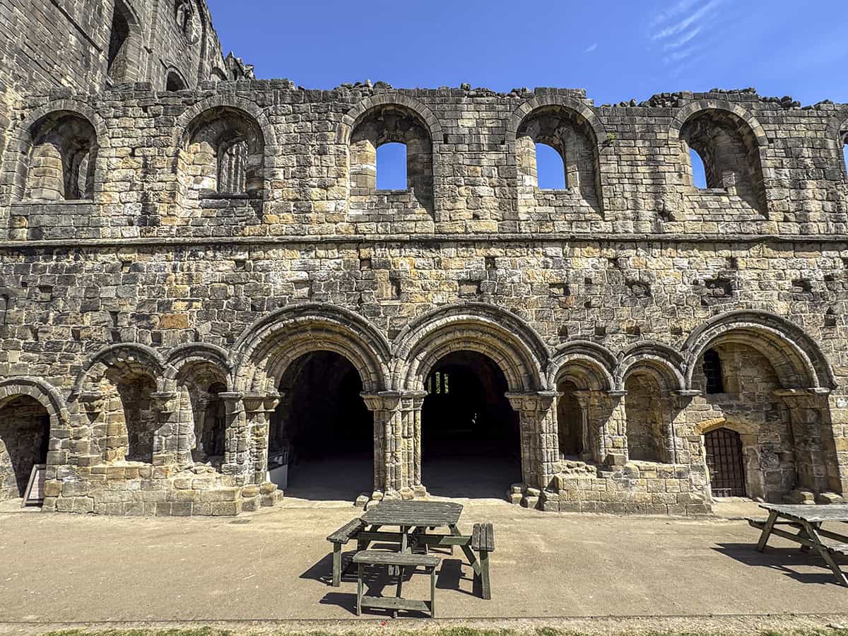 Exterior of Kirkstall Abbey Chapter House, with a double-arched entrance below a row of arched windows, and a tarmac path with picnic benches in front.