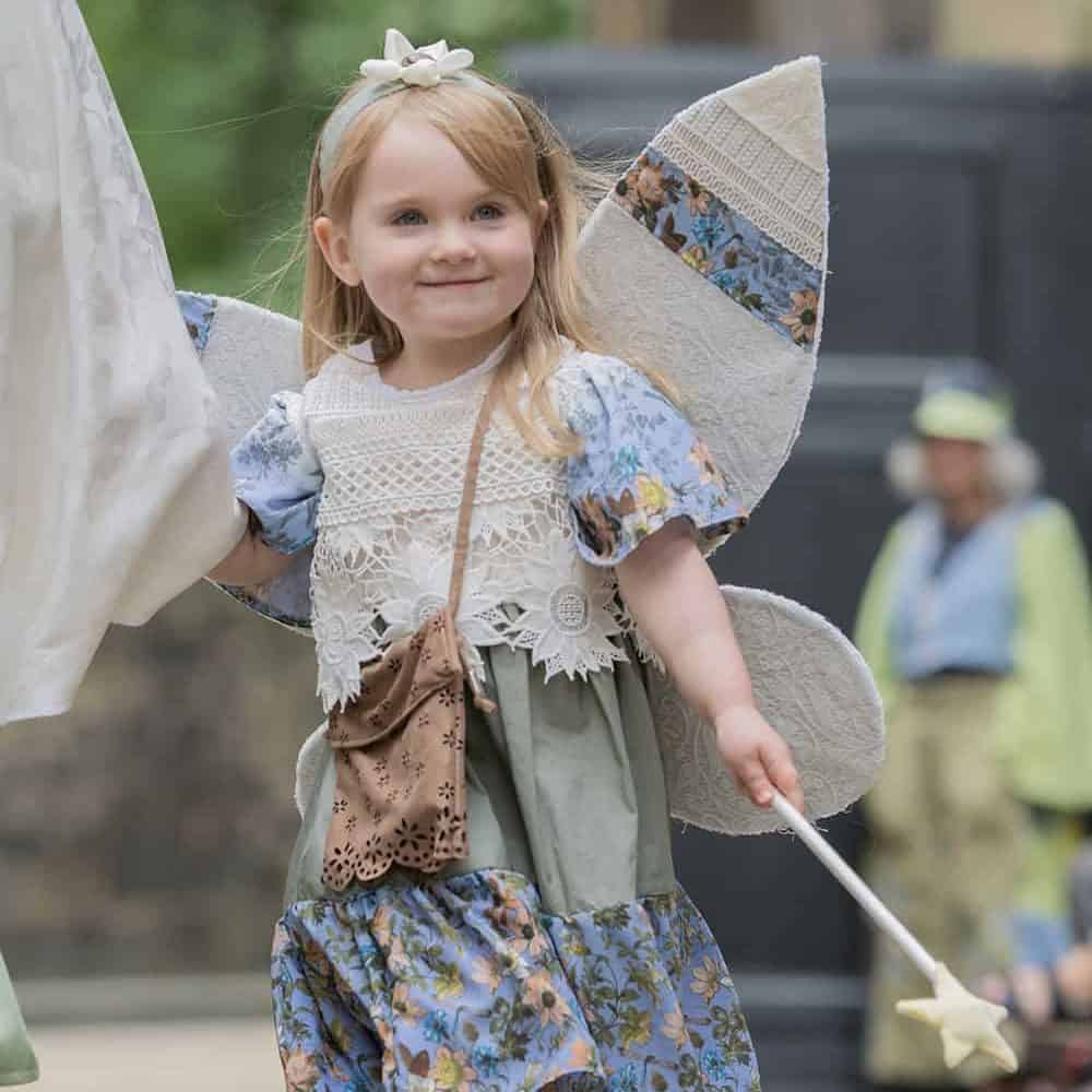 A young girl wears fairy wings and a matching dress, she carries a wand.