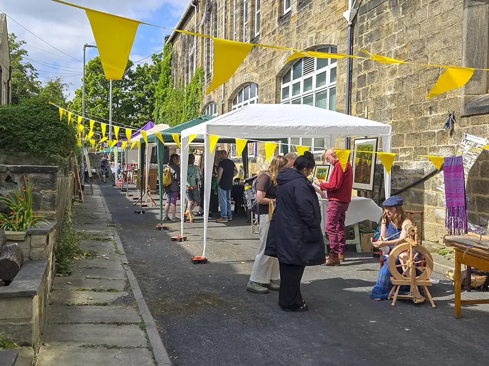Photograph of Victoria Terrace, a paved lane beside the rear of St Stephen's C of E Primary School, strung with yellow bunting. White and green gazebo stalls display artwork and craft items, with visitors browsing and a woman using a spinning wheel in the foreground.
