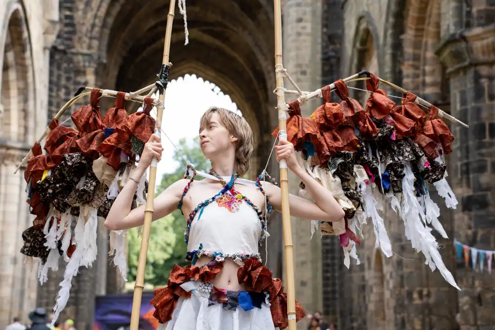 A model in a white halter top and skirt with rust-coloured fabric trim holds two tall poles hung with rust and cream fabric strips, resembling wings.