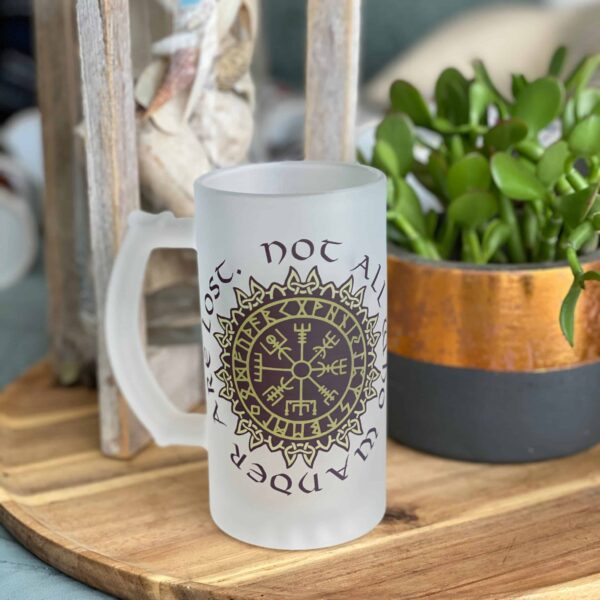 A Vegvisir Nor all who wander are lost frosted glass mug sits on a wooden tray next to a potted plant and a decorative glass container of seashells.