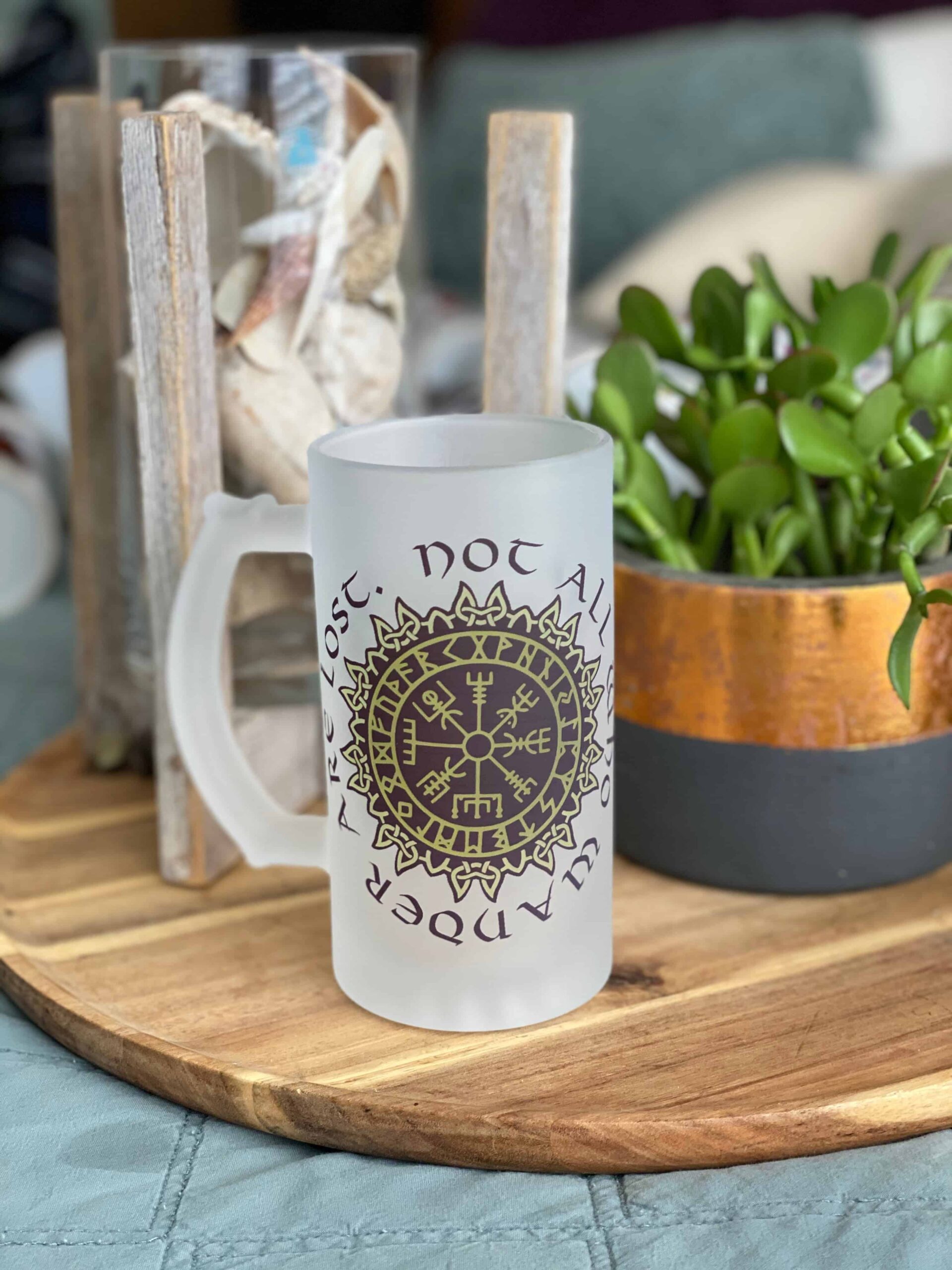 A Vegvisir Nor all who wander are lost frosted glass mug sits on a wooden tray next to a potted plant and a decorative glass container of seashells.