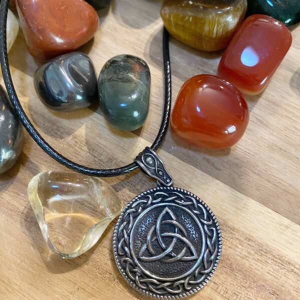 A Valknut Pendant Amulet Necklace with a Celtic knot design rests on a wooden surface, displayed on a black cord and surrounded by polished multicolored stones in red, green, yellow, and gray.