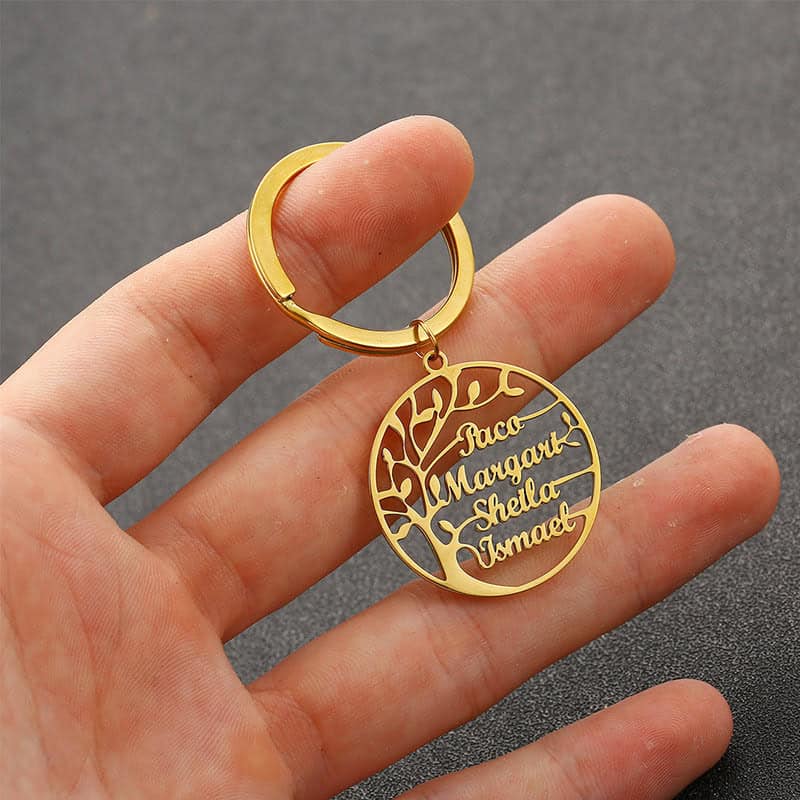 A hand holds a Custom Name Tree Of Life Stainless Steel Keychain, featuring a round gold design with a tree and the names Paco, Margaret, Sheila, and Ismael engraved in cursive on its branches.
