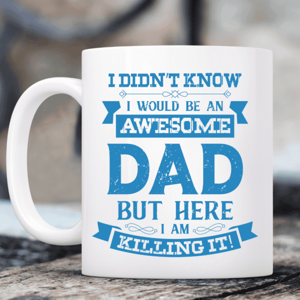 The IM An Awesome Dad Coffee Mug, white with blue text that reads “I didn’t know I would be an awesome dad but here I am killing it!”, rests on a table with a softly blurred background.