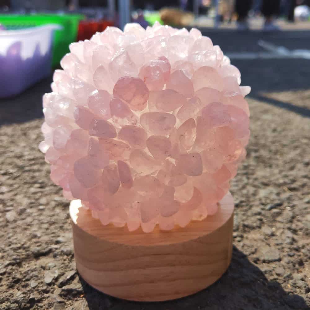 The Natural Crystal Ball Night Light features a cluster of pink crystals tightly arranged on a small wooden base, displayed outdoors on a textured surface with blurred containers in the background.