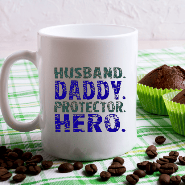 A white mug with bold blue and green text reading Husband Daddy Protector Hero sits on a green-checked cloth beside coffee beans and two chocolate muffins in green wrappers.