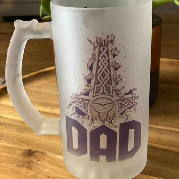 The Viking Thor’s Hammer Custom Beer Mug features a frosted glass design with a bold Thor’s Hammer above “DAD,” displayed on a wooden surface with a green plant in the background.