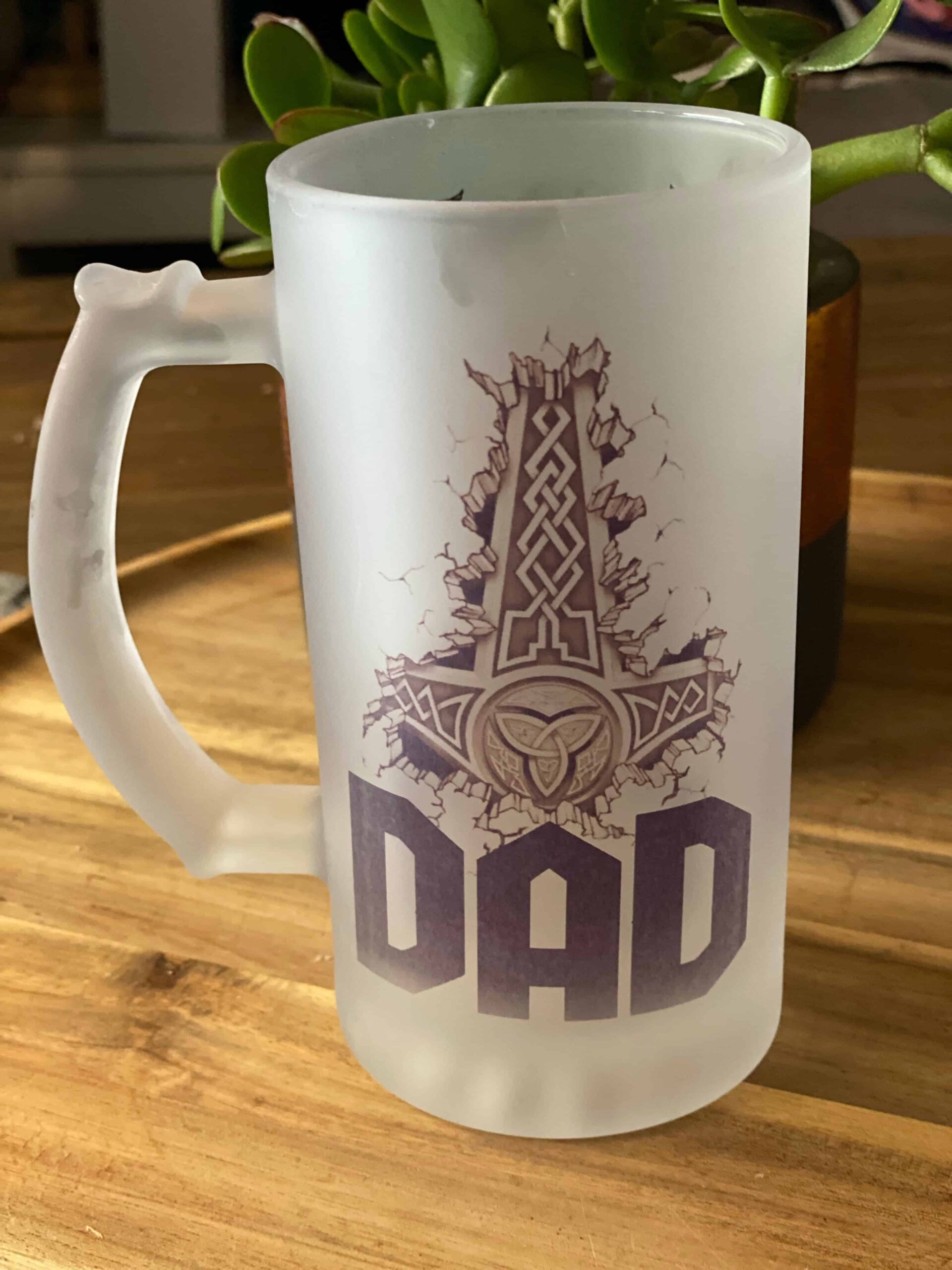 The Viking Thor’s Hammer Custom Beer Mug features a frosted glass design with a bold Thor’s Hammer above “DAD,” displayed on a wooden surface with a green plant in the background.