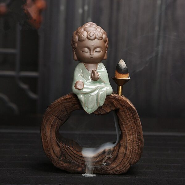 The Little Buddha Backflow Incense Burner features a small ceramic Buddha in green robes atop a wooden ring, with an incense cone beside it releasing smoke that gently cascades down through the ring.