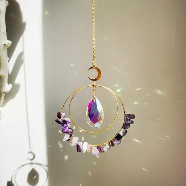 The Crystal Moon Suncatchers feature a crescent moon, faceted teardrop crystal, and semicircle of amethyst stones on a chain, casting rainbow reflections when sunlight shines through.