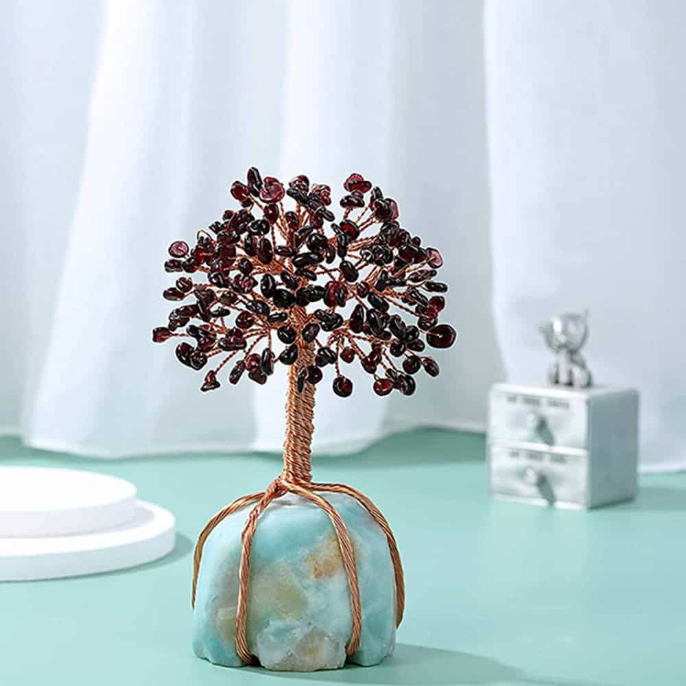 A Natural Gem Stone & Copper Feng Shui Tree with wire branches and red gemstone leaves on a blue stone base is displayed on a light green surface, with a white curtain and small silver ornament in the background.