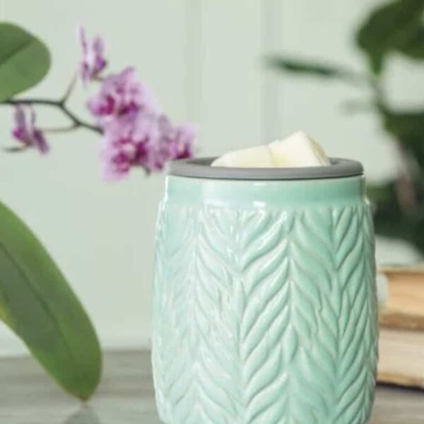 The Mint Flip Dish Wax Warmer, featuring a leaf pattern, holds white wax melts on a table. A softly blurred purple flower and green leaves appear in the background.