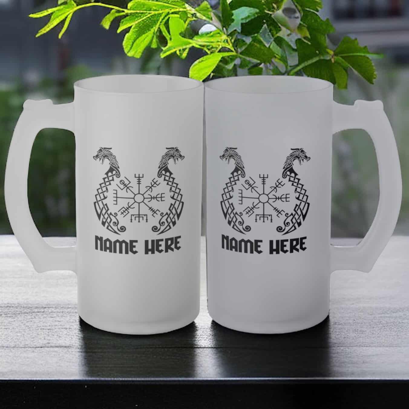 Two Viking Dragon Boat Frosted Glass Beer Mug Personalised rest on a dark table, each with dragon designs, runic symbols, and NAME HERE below. A green plant and an outdoor background complete the scene.