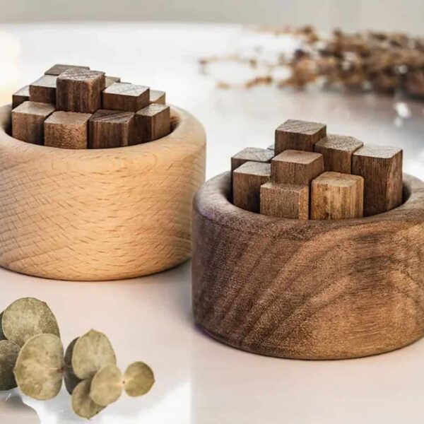 Two round wooden containers, one light and one dark, each hold square wooden blocks in rows. They sit on a white surface alongside the Wooden Essential Oil Diffuser, leaves, and other decorative items.