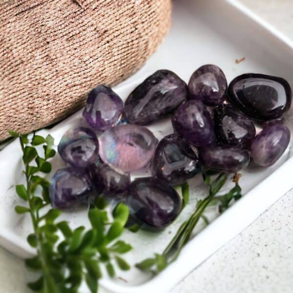A white tray displays several small Amethyst Tumble Stones, complemented by green leafy sprigs and a hint of textured pink fabric in the background.