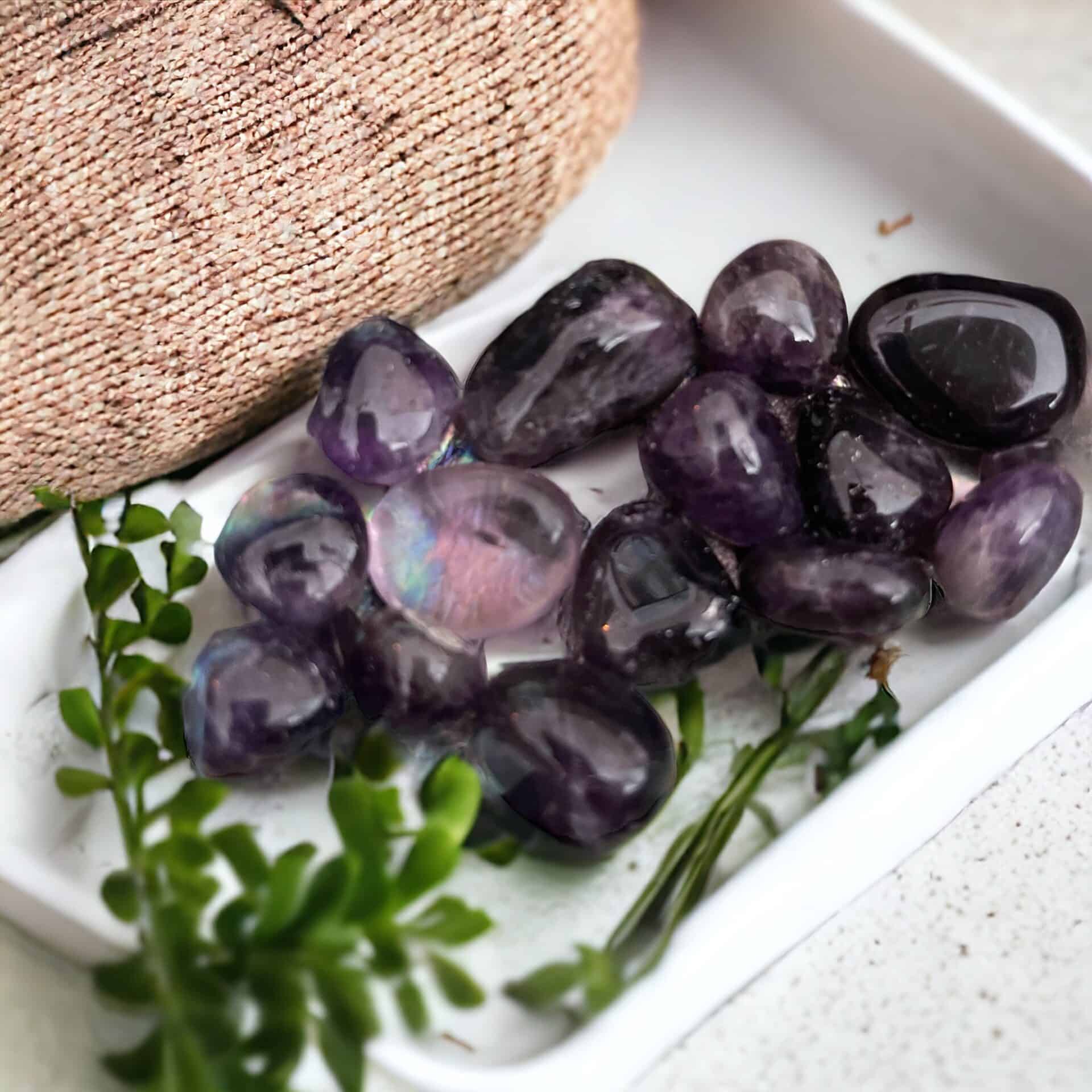A white tray displays several small Amethyst Tumble Stones, complemented by green leafy sprigs and a hint of textured pink fabric in the background.