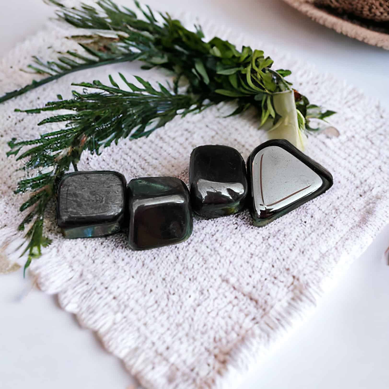 Four Hematite Tumble Stones are displayed on a textured white mat with green foliage, creating a calm and natural scene.