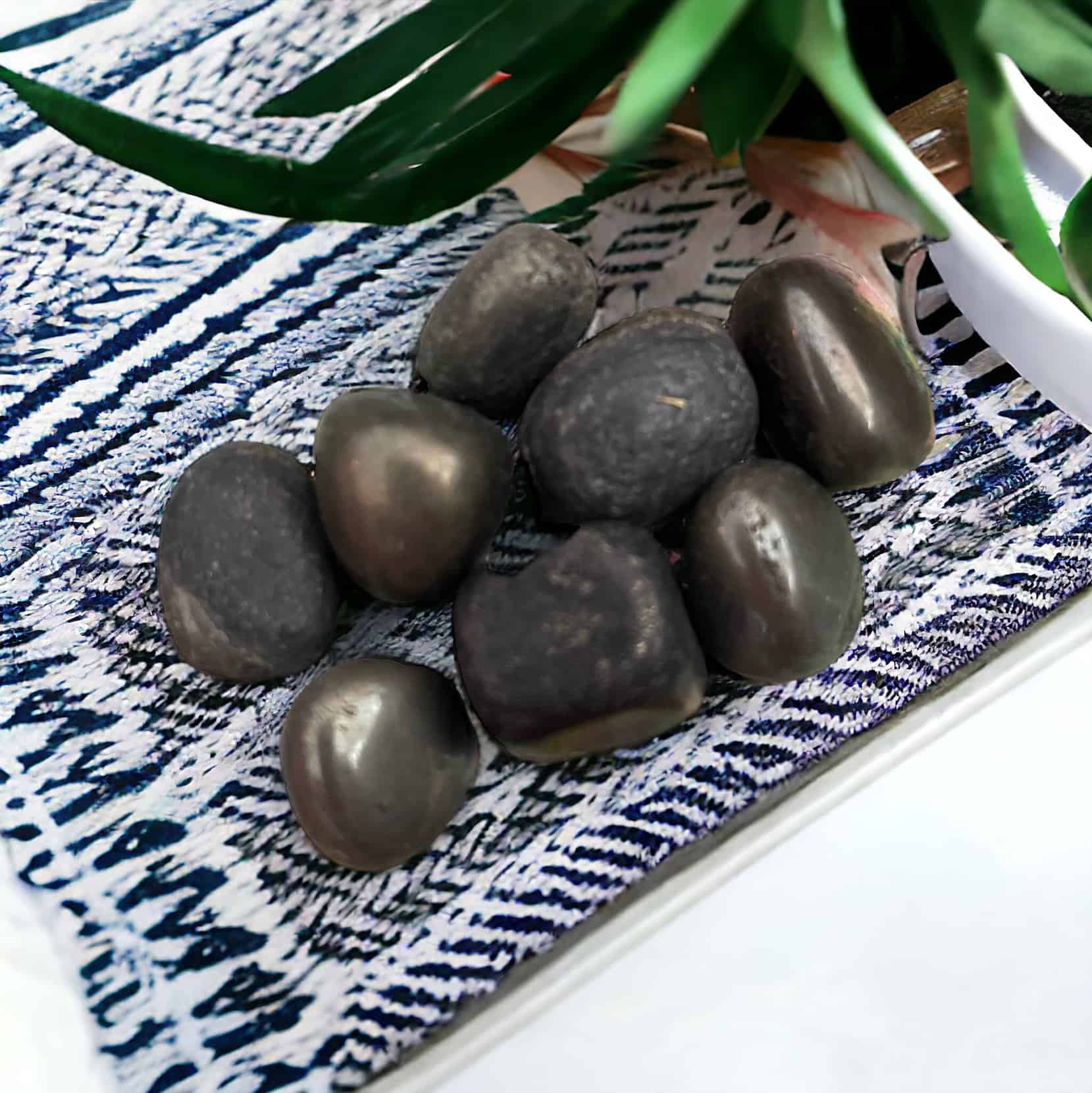 A small pile of shiny, dark Pyrite Tumble Stones rests on patterned blue and white fabric, with green leaves from a potted plant visible in the background.