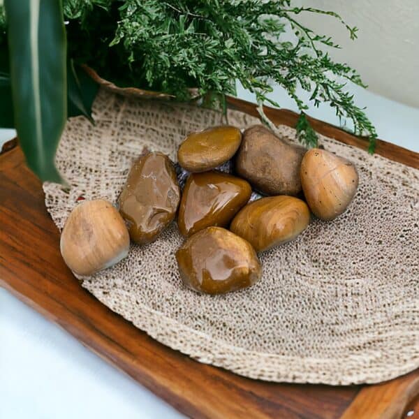 A group of Yellow Jasper Tumble Stones are arranged on a woven beige mat atop a wooden tray, with green foliage in the background.