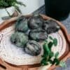 A wooden tray displays six polished Emerald Tumble Stone Crystals, their rich green and black hues gleaming atop a woven mat, surrounded by lush potted plants and greenery.