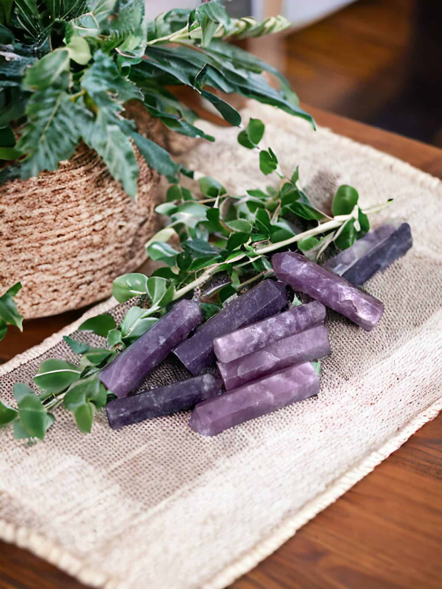 A Lepidolite Mini Tower Crystal, polished to a shine, sits with other purple stones on a burlap cloth accented by green leaves. In the background, a woven basket with a leafy plant rests on a wooden surface.
