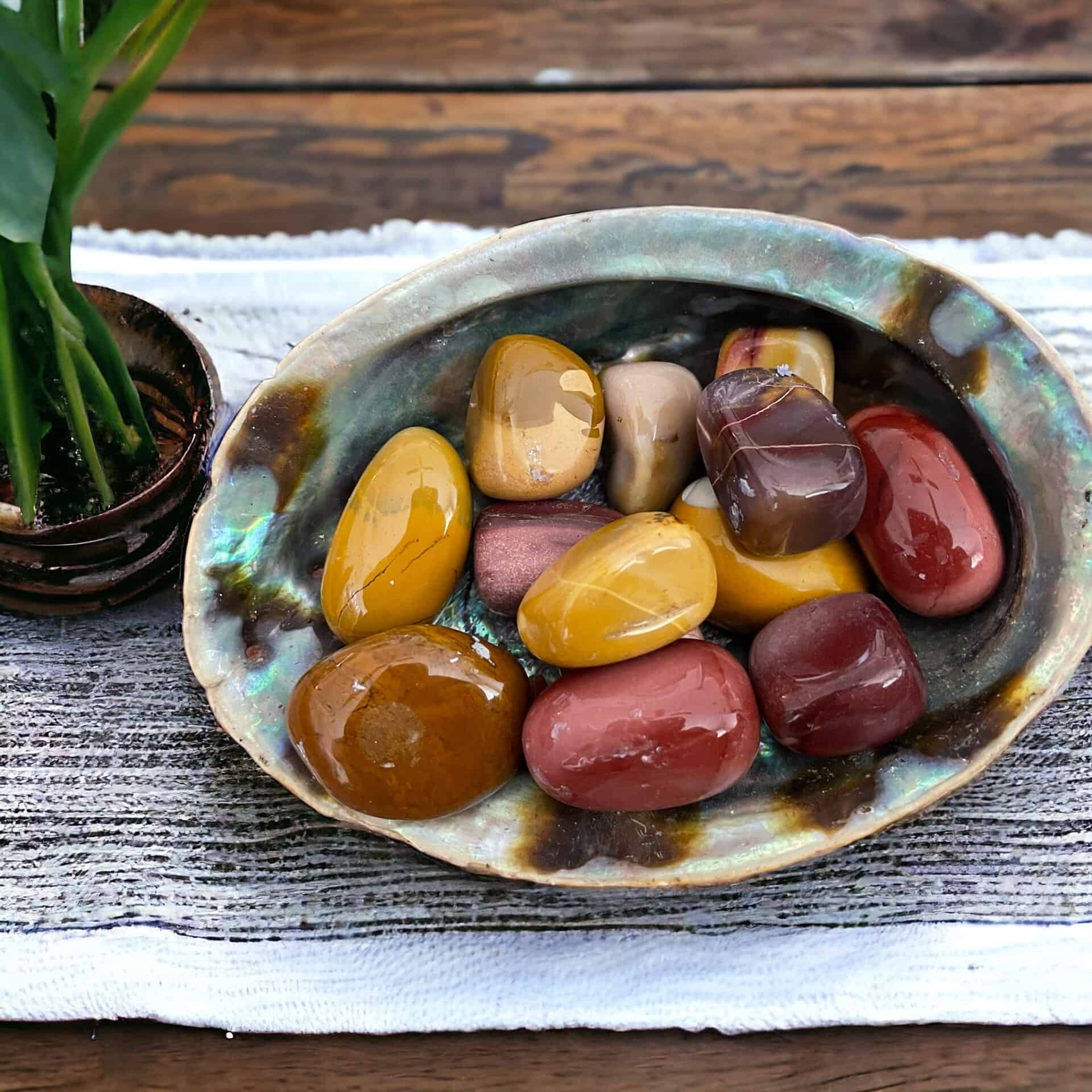 A ceramic bowl filled with Mookaite Tumble Stones in yellow, red, brown, and tan rests on a woven cloth beside a small potted plant atop a wooden table.