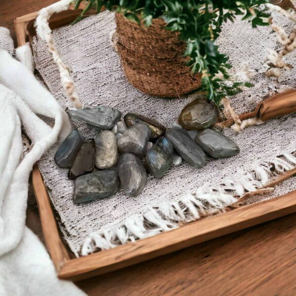 A wooden tray holds a plant wrapped in twine, several Labradorite Tumble Stones, and a fringed woven cloth, all set atop wood beside soft white fabric.