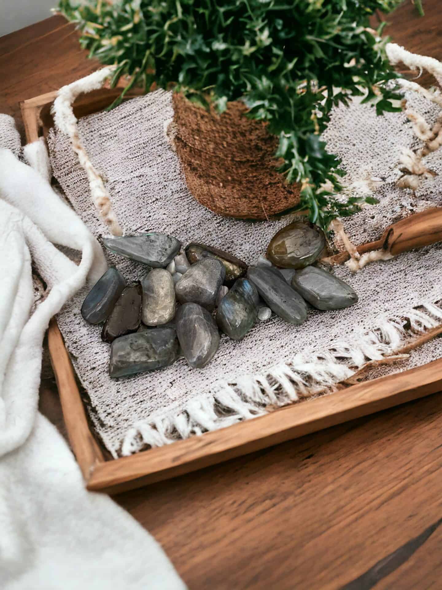A wooden tray holds a plant wrapped in twine, several Labradorite Tumble Stones, and a fringed woven cloth, all set atop wood beside soft white fabric.