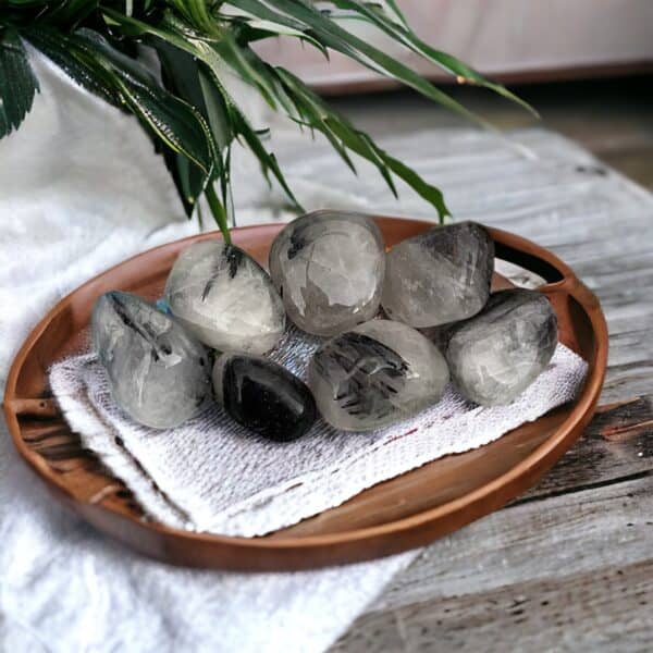 Tourmalated Quartz—polished stones with black streaks—rests on a wooden tray over a white cloth on rustic wood, with green plant leaves arching above.