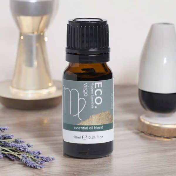 A bottle of Virgo Essential Oil Blend rests on a table beside lavender, an hourglass, and decor. The green label with white and gold accents identifies it as the Virgo Essential Oil Blend.