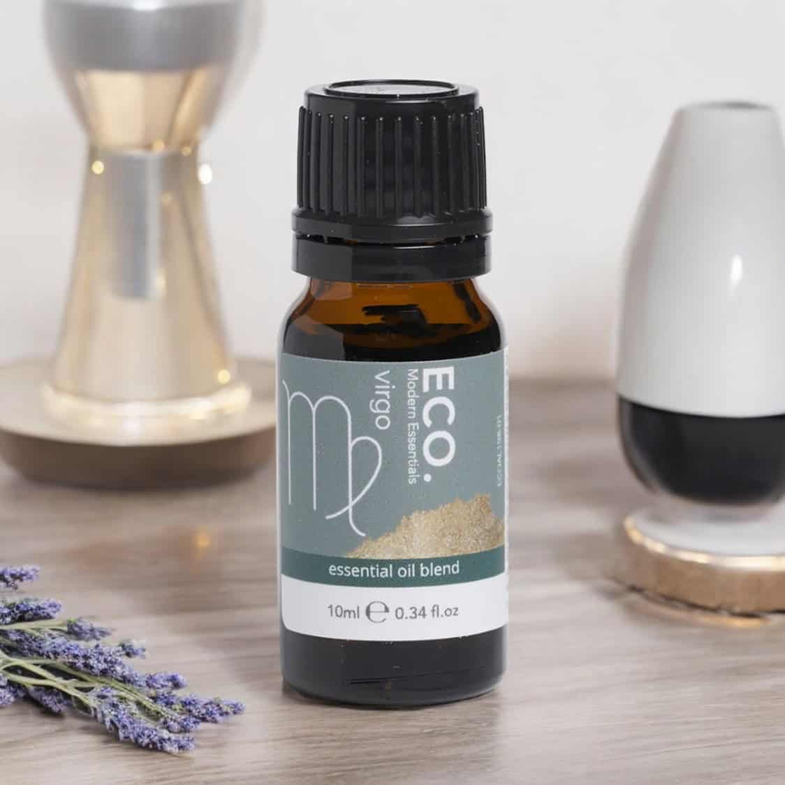 A bottle of Virgo Essential Oil Blend rests on a table beside lavender, an hourglass, and decor. The green label with white and gold accents identifies it as the Virgo Essential Oil Blend.