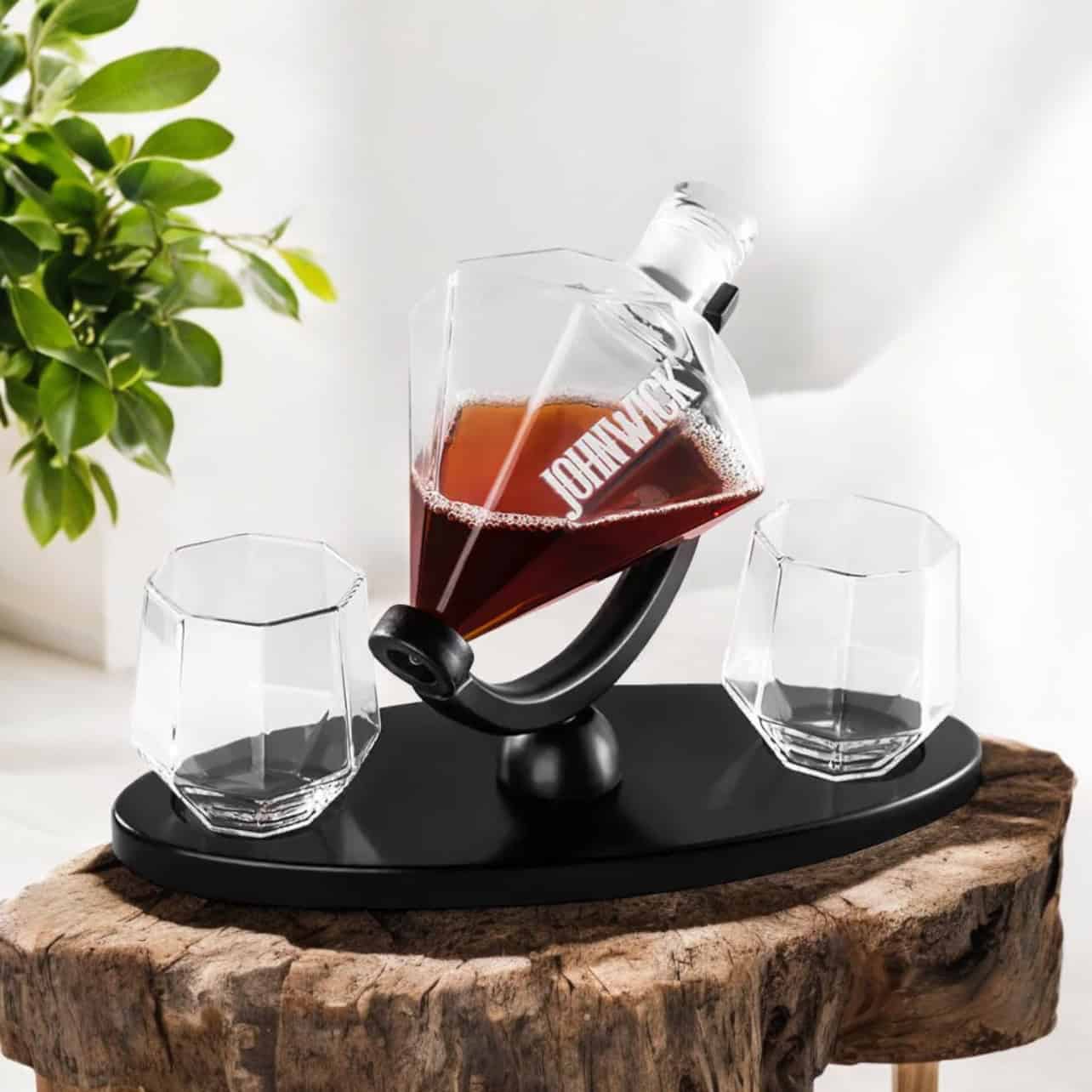The Engraved Decanter and Glass Set features a modern decanter on a black stand, paired with two matching glasses, all displayed on a rustic wooden surface beside a green plant.