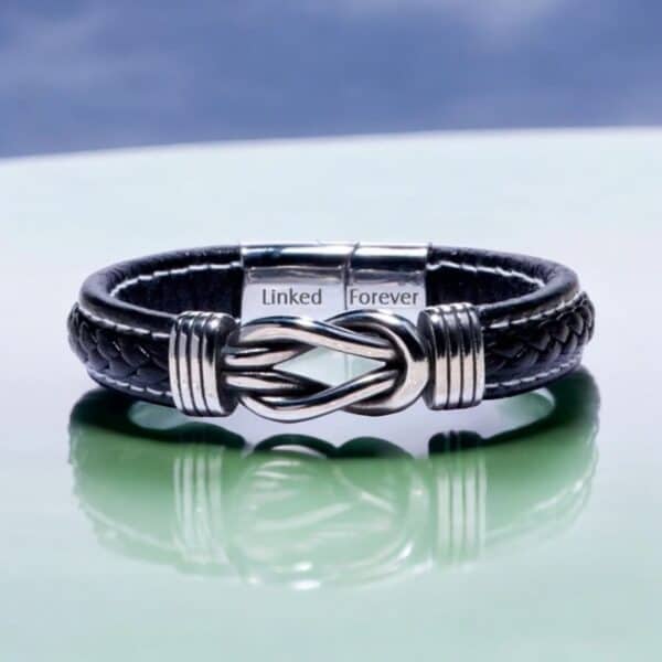 The Men’s Personalised Link Bracelet features black braided leather, a silver clasp with a knot design, and is engraved with Linked Forever. Its showcased on a glossy pale surface with a softly blurred blue and purple background.