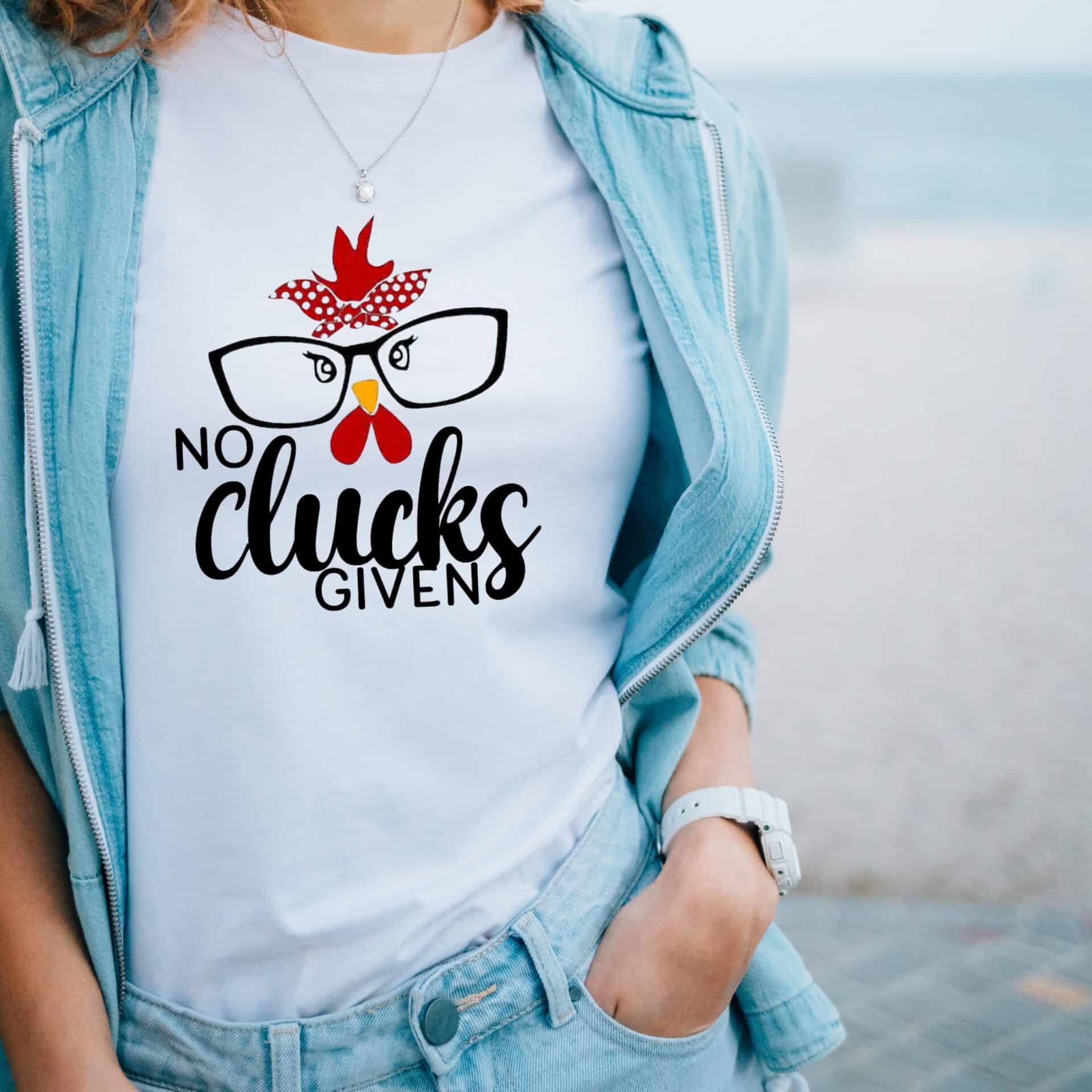 A woman stands outdoors in a light blue jacket and jeans, wearing the No Clucks Given Women’s Tee featuring a cartoon chicken face, red bandana, glasses, and the text “No Clucks Given” on a white Womens T-Shirt.