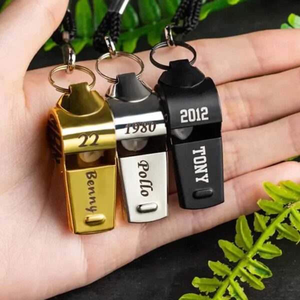 A hand holds three Personalised Whistle Necklaces—gold, silver, and black—each engraved with unique names and numbers. Green fern leaves are visible at the bottom of the image.