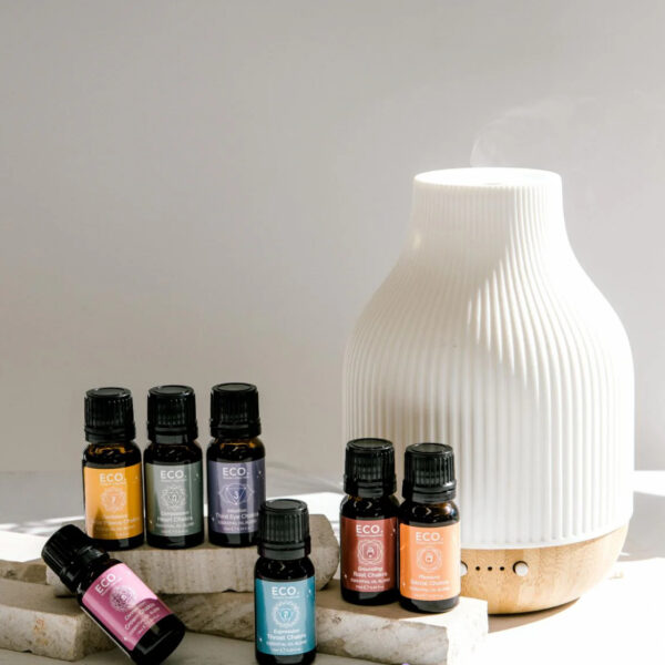 The Chakra Collection with Diffuser Aromatherapy Bundle features a white ceramic diffuser and seven colorful essential oil bottles, elegantly arranged on stone blocks with steam rising, all set against a neutral background.