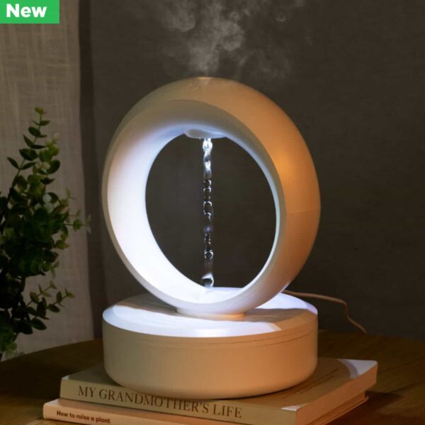 The Eclipse Anti-Gravity Diffuser features water flowing vertically inside a modern white ring. Placed on books beside a small potted plant, soft lighting enhances its calming ambiance.