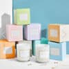 Colorful pastel boxes of Hidden Jewels Candle are stacked on a white surface, with two white glass jar candles in front. Small pieces of jewelry surround the candles, set against a blue and white background.