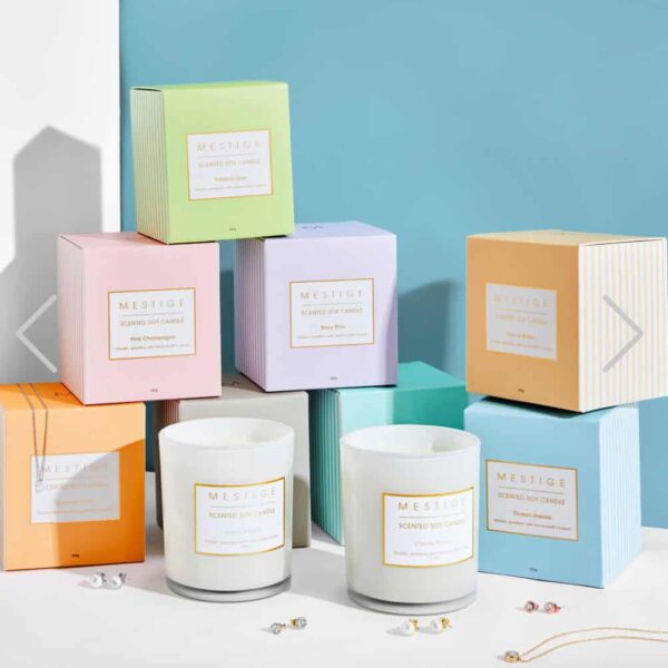 Colorful pastel boxes of Hidden Jewels Candle are stacked on a white surface, with two white glass jar candles in front. Small pieces of jewelry surround the candles, set against a blue and white background.