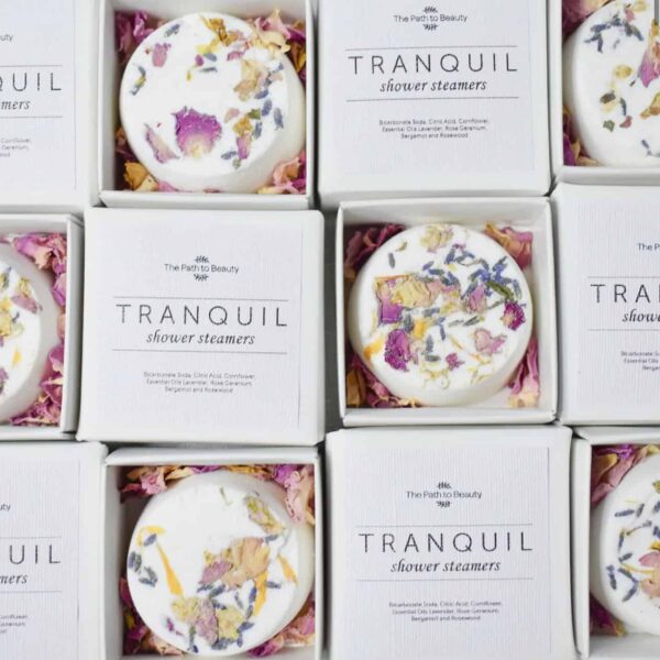Open white boxes of Tranquil Individual Shower Steamers, each topped with pink and yellow dried flower petals. Arranged in a grid, the packaging reveals both the shower steamer and its product label inside.