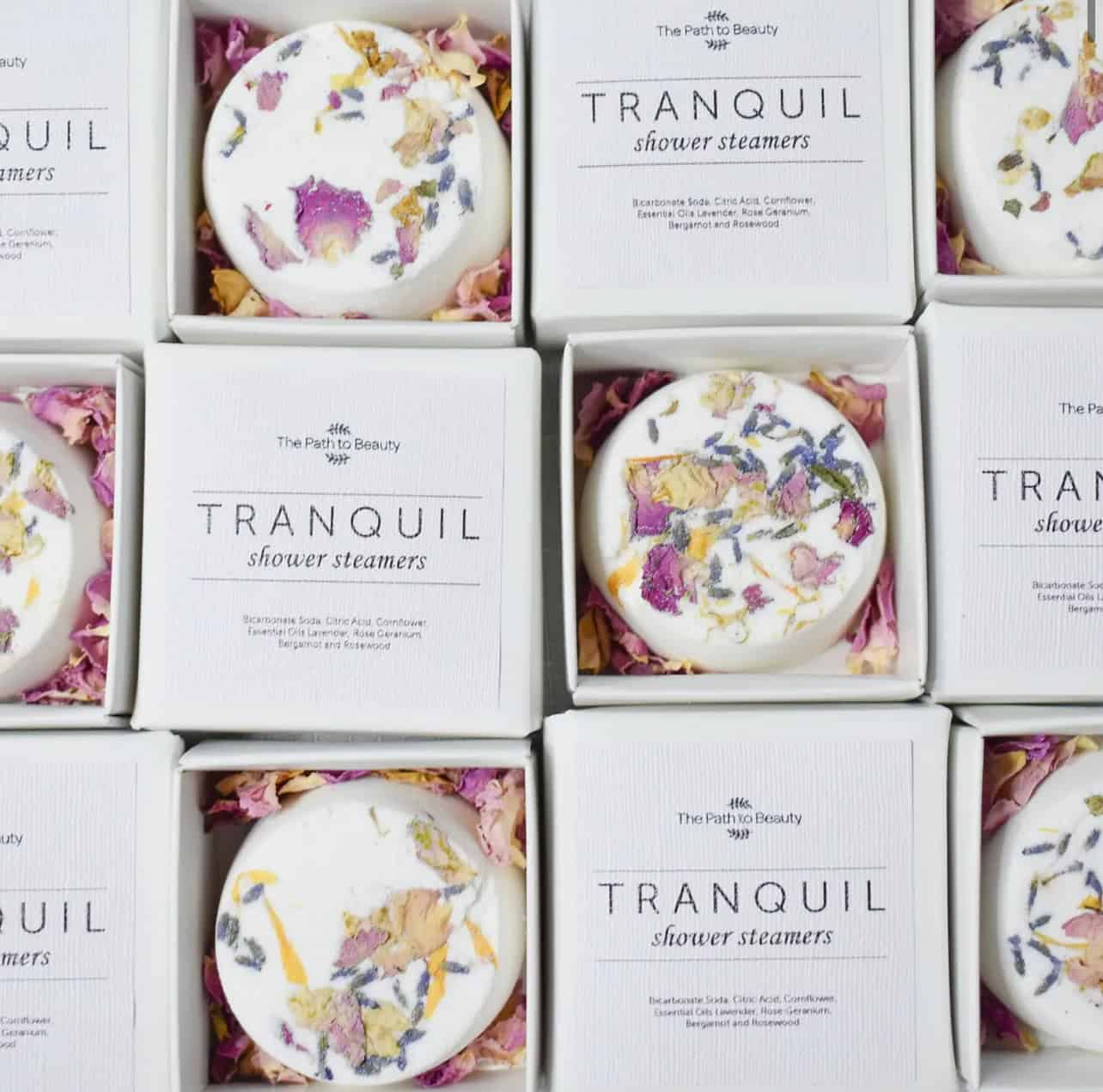 Open white boxes of Tranquil Individual Shower Steamers, each topped with pink and yellow dried flower petals. Arranged in a grid, the packaging reveals both the shower steamer and its product label inside.