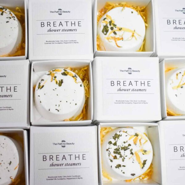 Nine boxes of Breathe Individual Shower Steamers are arranged in a grid. Each round, white steamer with dried herbs rests in yellow shredded paper inside its labeled box.